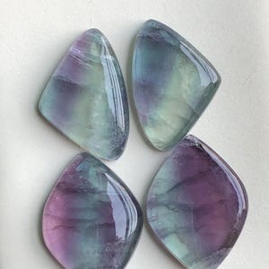 May include: Four polished fluorite gemstones. Each stone has a unique shape, with a mix of purple, green, and blue hues. The stones have a glossy, reflective surface, and are arranged on a white background.
