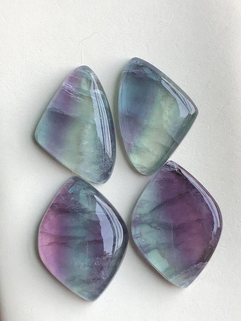 May include: Four polished fluorite gemstones. Each stone has a unique shape, with a mix of purple, green, and blue hues. The stones have a glossy, reflective surface, and are arranged on a white background.