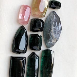 K&ouml;nnte beinhalten: Eine Sammlung von zehn facettierten Edelsteinen in verschiedenen Formen und Gr&ouml;&szlig;en. Die Steine zeigen Schattierungen von tiefem Schwarz, dunklem Waldgr&uuml;n, durchscheinendem Rosa und blassem Gelbgr&uuml;n sowie einen gro&szlig;en, l&auml;nglichen, graublauen Stein.