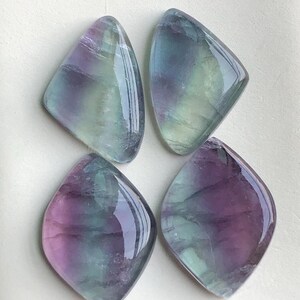 May include: Four polished fluorite gemstones. Each stone is a unique shape, with a mix of purple, green, and clear colours. The stones have a glossy, reflective surface. The gemstones are arranged on a white surface.