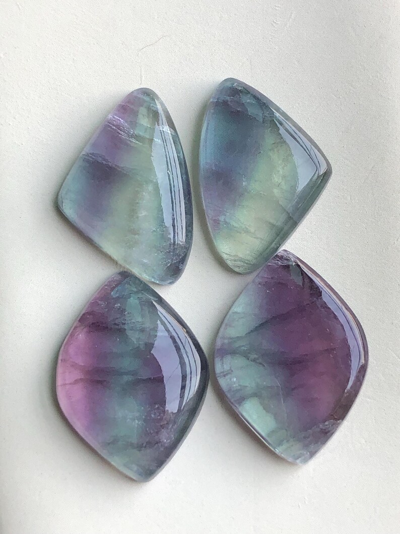 May include: Four polished fluorite gemstones. Each stone is a unique shape, with a mix of purple, green, and clear colours. The stones have a glossy, reflective surface. The gemstones are arranged on a white surface.