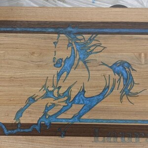 May include: A wooden cutting board with a blue resin inlay of a horse design. The board has a walnut border and the name "Laura" is engraved on the bottom.