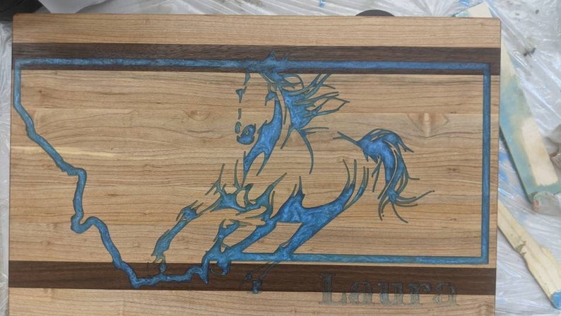 May include: A wooden cutting board with a blue resin inlay of a horse design. The board has a walnut border and the name "Laura" is engraved on the bottom.
