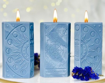 Puede incluir: Tres velas azules con diseños de cartas de tarot. Las velas están encendidas y tienen un fondo blanco. La vela del medio tiene el diseño de la carta de la Rueda de la Fortuna.