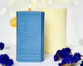 Puede incluir: Una vela azul con un diseño en relieve de la carta del As de Pentáculos del tarot. La vela está encendida y tiene una llama amarilla.