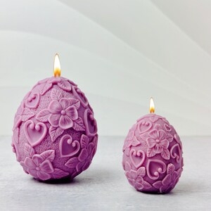 May include: Two purple candles shaped like eggs with floral and heart designs. The larger candle is lit.