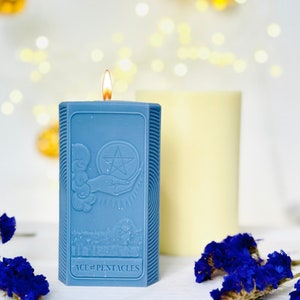 May include: A blue candle featuring a raised design of the Ace of Pentacles tarot card. The candle is lit and has a yellow flame.