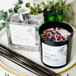 May include: A black candle and a clear diffuser with the text "LOVE SPELL" and "infused with Rose Quartz + Amethyst". The candle is filled with crystals and flowers. The diffuser has a silver top. The items are on a white plate with a gold rim.