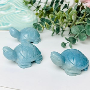 May include: Three blue-green turtle figurines carved from stone. The turtles are arranged in a row on a white surface.