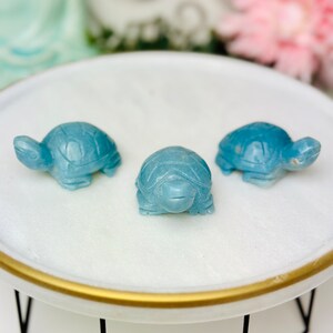 May include: Three small blue turtle figurines on a white round plate with a gold rim. The plate is on a black metal stand.