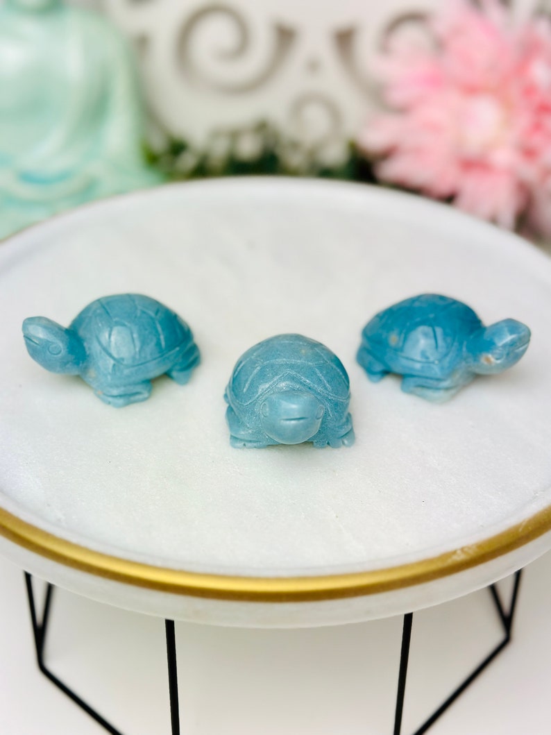 May include: Three small blue turtle figurines on a white round plate with a gold rim. The plate is on a black metal stand.