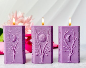 May include: Three purple pillar candles with a tree and moon design. The candles are on a white surface.