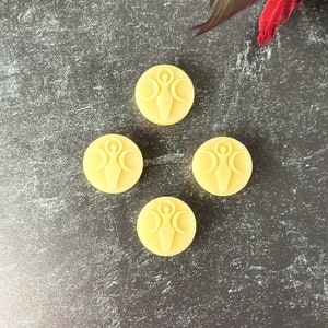 May include: Four round, pale yellow wax melts with a raised design of a woman standing between three crescent moons.