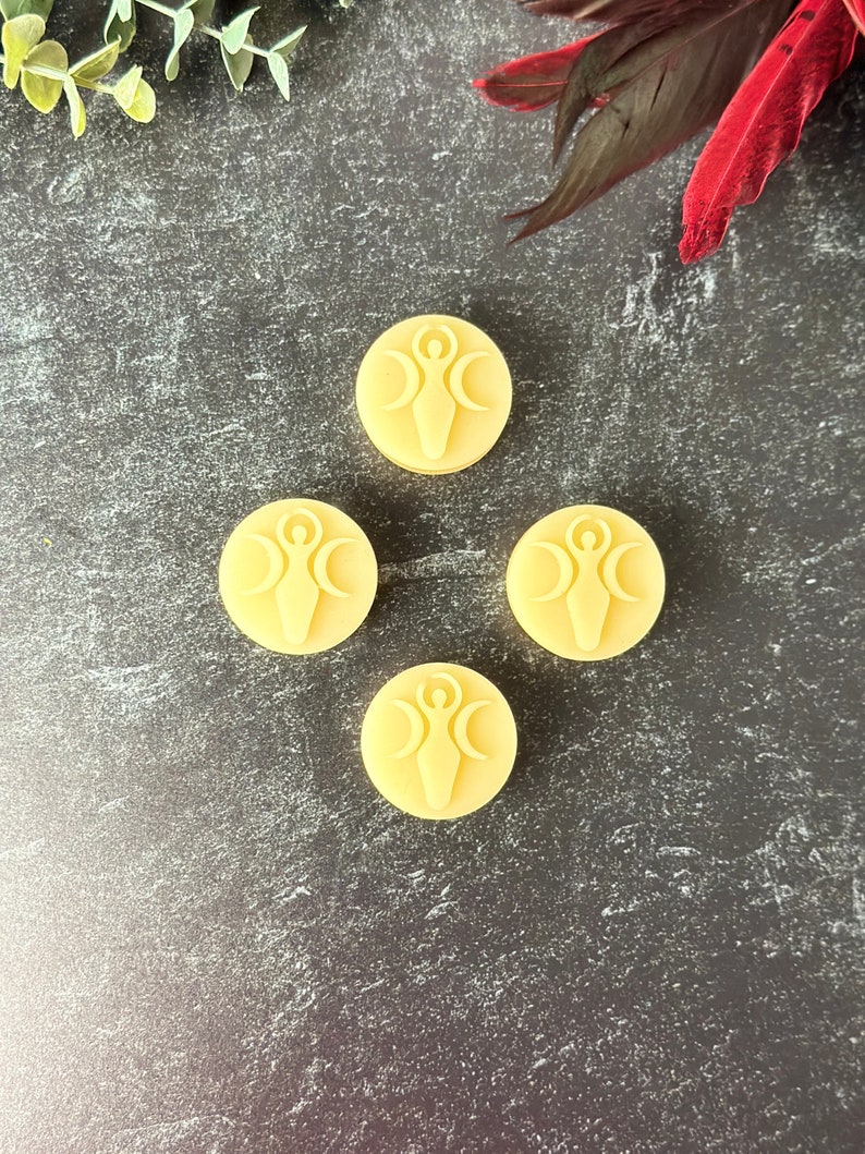 May include: Four round, pale yellow wax melts with a raised design of a woman standing between three crescent moons.