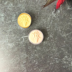 May include: Two small, round, votive candles with a raised design of a woman with outstretched arms and a crescent moon above her head. The candles are on a dark gray textured surface. One candle is yellow and the other is peach.