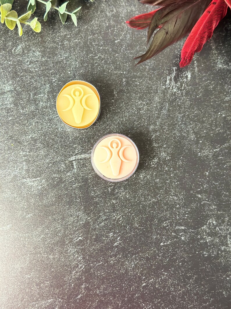 May include: Two small, round, votive candles with a raised design of a woman with outstretched arms and a crescent moon above her head. The candles are on a dark gray textured surface. One candle is yellow and the other is peach.