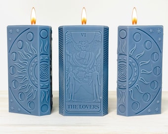 May include: Three blue candles with a tarot card design. The middle candle features the Lovers card from the tarot deck. The candles are decorated with a sun and moon design.