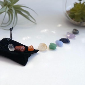May include: A set of seven chakra stones in a black velvet pouch. The stones are arranged in a semi-circle, with the colors of the chakras: red, orange, yellow, green, blue, indigo, and violet.