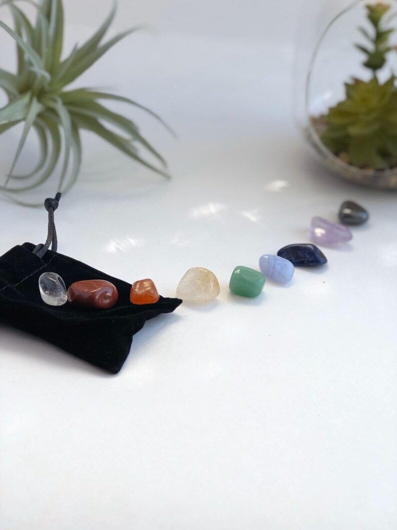 May include: A set of seven chakra stones in a black velvet pouch. The stones are arranged in a semi-circle, with the colors of the chakras: red, orange, yellow, green, blue, indigo, and violet.