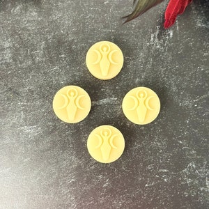 May include: Four round, pale yellow wax melts with a raised design of a woman standing with three crescent moons above her head. The wax melts are arranged in a diamond shape on a dark gray textured surface.