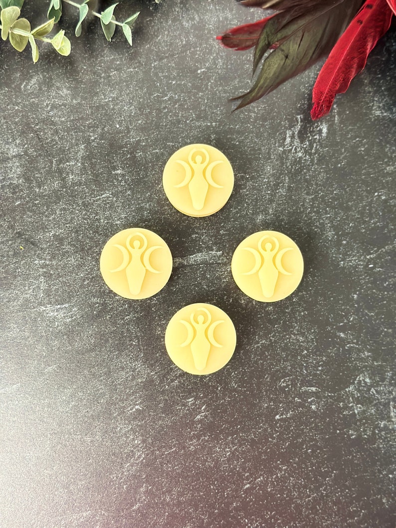 May include: Four round, pale yellow wax melts with a raised design of a woman standing with three crescent moons above her head. The wax melts are arranged in a diamond shape on a dark gray textured surface.