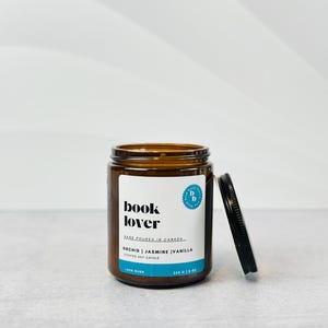 May include: A brown glass jar candle with a black lid. The label reads "book lover" and "orchid | jasmine | vanilla scented soy candle". The jar is 350 g or 12 oz.