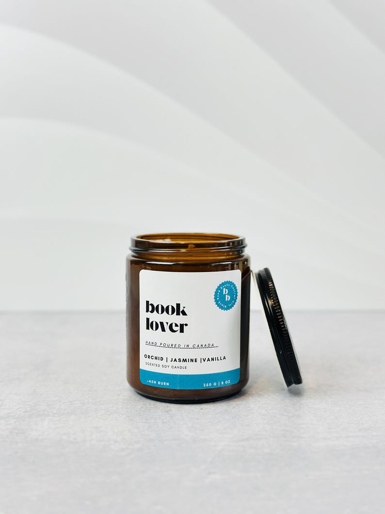 May include: A brown glass jar candle with a black lid. The label reads "book lover" and "orchid | jasmine | vanilla scented soy candle". The jar is 350 g or 12 oz.
