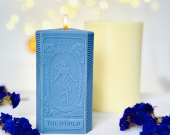 Puede incluir: Una vela azul con un diseño de carta de tarot. La vela está encendida y el diseño presenta a una mujer de pie en un círculo con el número XXI y las palabras "THE WORLD" en la parte inferior.