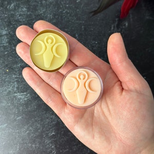 May include: Two small, round, wax melts in clear containers. The wax melts are in the shape of a circle with a woman symbol in the center. One wax melt is yellow and the other is peach.