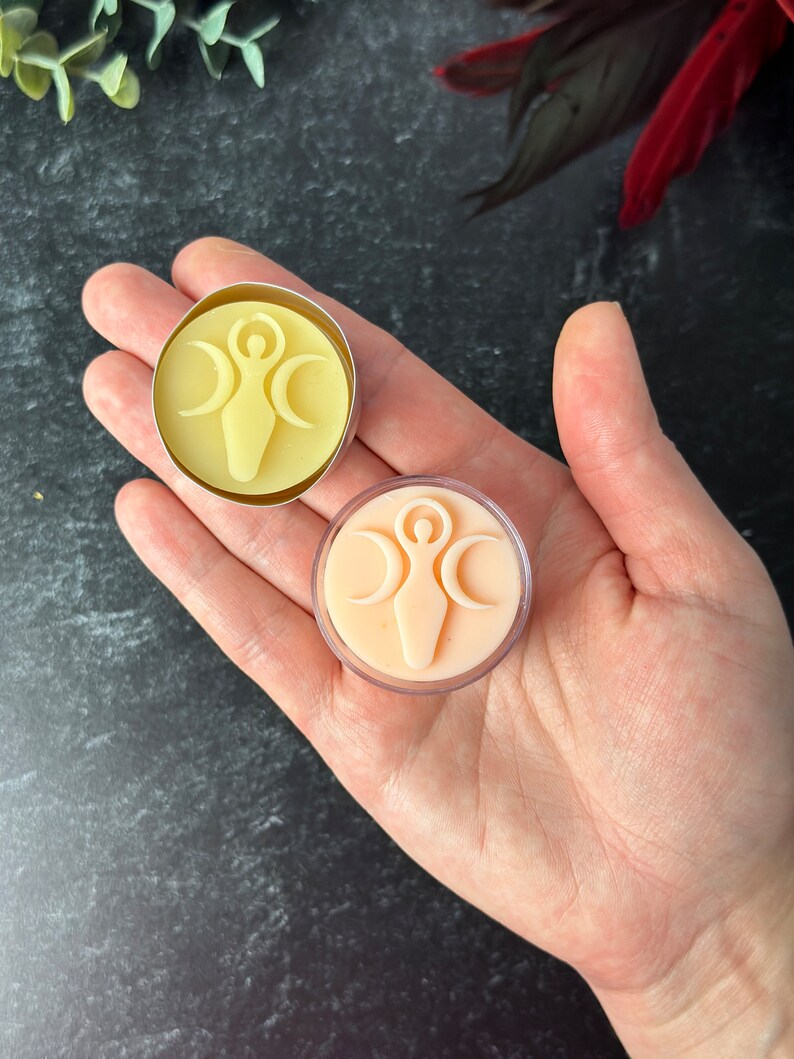 May include: Two small, round, wax melts in clear containers. The wax melts are in the shape of a circle with a woman symbol in the center. One wax melt is yellow and the other is peach.