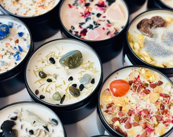 May include: Assortment of small, round candles in black tins. Each candle has a different combination of crystals, herbs, and dried flowers embedded in the wax. The candles are arranged in a circular pattern on a white surface.