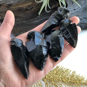 May include: A collection of six black obsidian arrowhead points. The arrowheads are smooth and polished, with sharp edges. They are arranged in a fan shape on a hand.