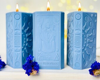 Puede incluir: Tres velas azules de tarot con un diseño de la carta del Hierofante, un diseño de sol y luna y un diseño de sol y luna. Las velas están encendidas y tienen un brillo cálido.