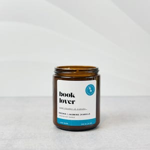 May include: A brown glass jar candle with a white label that reads "book lover" and "orchid | jasmine | vanilla scented soy candle". The label also includes the text "hand poured in canada" and a small blue circle with the letters "b b" inside.