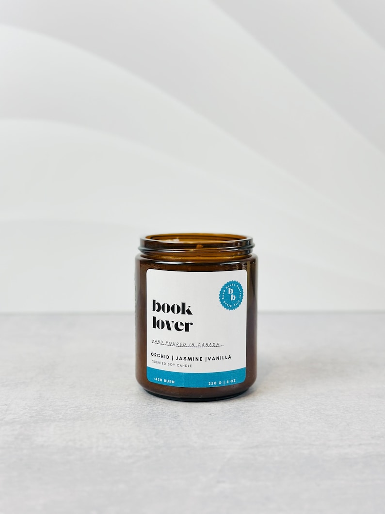 May include: A brown glass jar candle with a white label that reads "book lover" and "orchid | jasmine | vanilla scented soy candle". The label also includes the text "hand poured in canada" and a small blue circle with the letters "b b" inside.