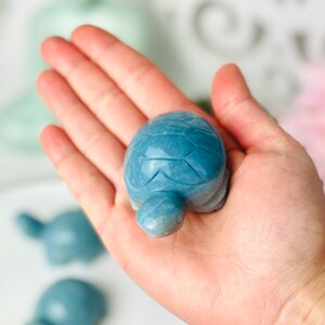 May include: A blue stone turtle figurine. The turtle is carved with details on its shell and head. The turtle is sitting on a white surface.