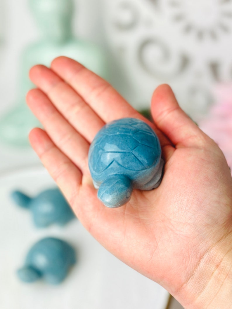 May include: A blue stone turtle figurine. The turtle is carved with details on its shell and head. The turtle is sitting on a white surface.