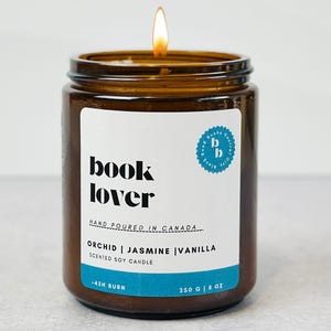 May include: A brown glass candle jar with a white label that reads "book lover" and "orchid | jasmine | vanilla scented soy candle". The candle is lit and the flame is visible.
