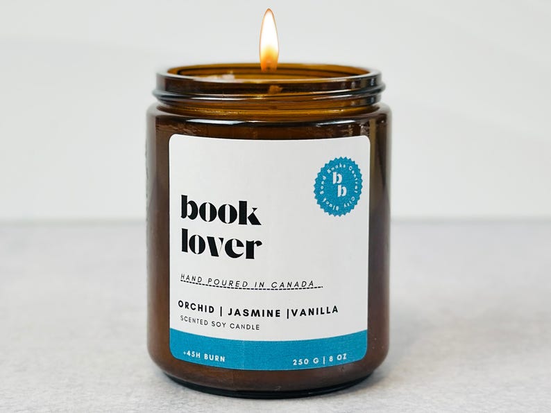 May include: A brown glass candle jar with a white label that reads "book lover" and "orchid | jasmine | vanilla scented soy candle". The candle is lit and the flame is visible.
