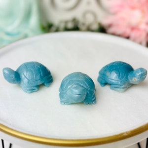 May include: Three small blue turtle figurines made of polished stone. The turtles are arranged in a row on a white circular surface with a gold rim.