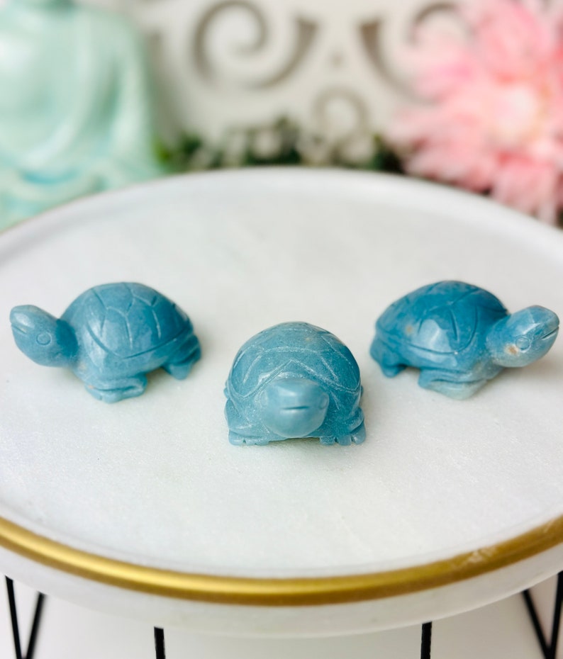 May include: Three small blue turtle figurines made of polished stone. The turtles are arranged in a row on a white circular surface with a gold rim.