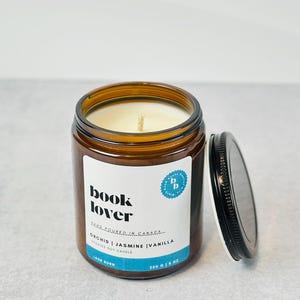 May include: A brown glass jar candle with a black lid. The candle is labeled "book lover" and "orchid | jasmine | vanilla scented soy candle".