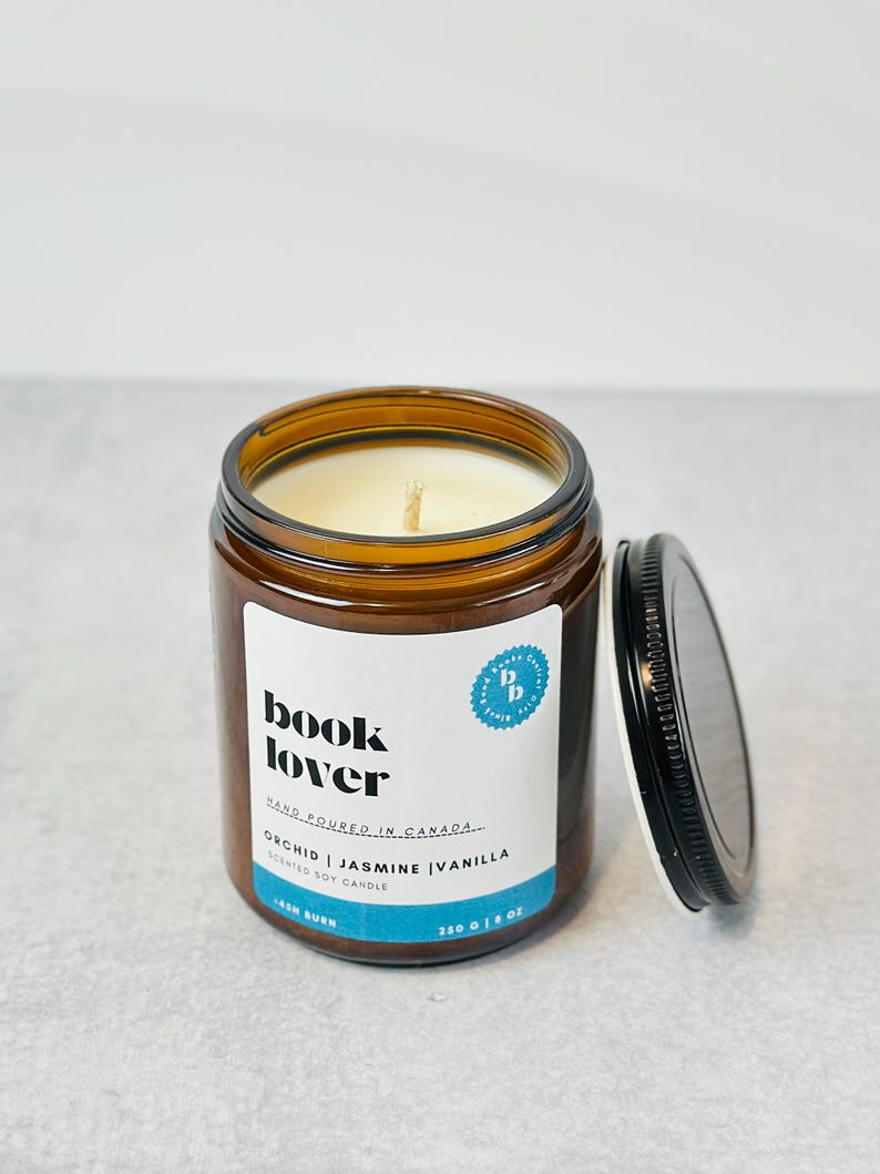 May include: A brown glass jar candle with a black lid. The candle is labeled "book lover" and "orchid | jasmine | vanilla scented soy candle".