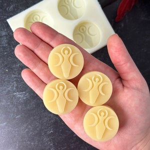 May include: Four round, pale yellow, wax melts with a raised design of a woman standing with her arms raised and three crescent moons around her.