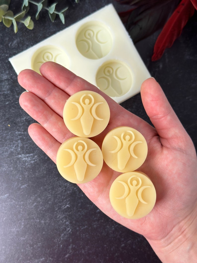 May include: Four round, pale yellow, wax melts with a raised design of a woman standing with her arms raised and three crescent moons around her.