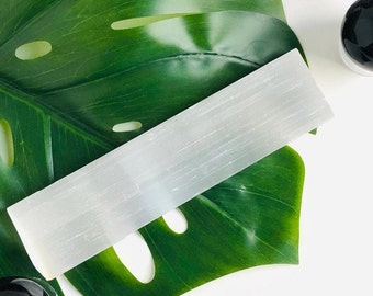 May include: A white selenite wand resting on a green monstera leaf. The wand is smooth and has a natural, striated texture.