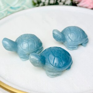 May include: Three blue turtle figurines made of polished stone. The turtles are arranged on a white circular surface with a gold rim.
