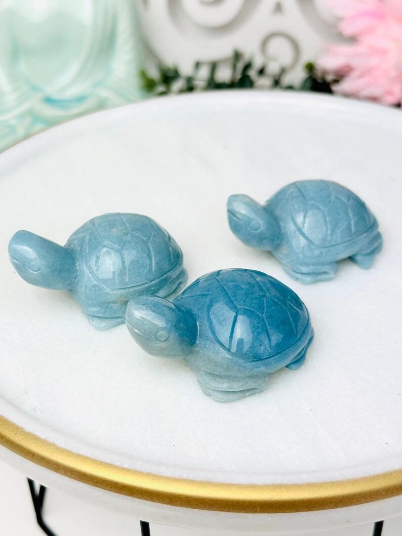 May include: Three blue turtle figurines made of polished stone. The turtles are arranged on a white circular surface with a gold rim.