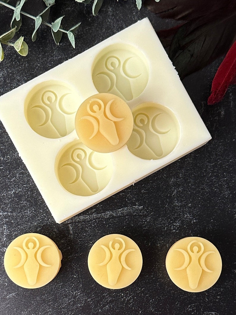 May include: A silicone mold with five circular impressions, each featuring a raised design of a woman standing with three crescent moons. There are four yellow wax melts with the same design, one of which is sitting on top of the mold.