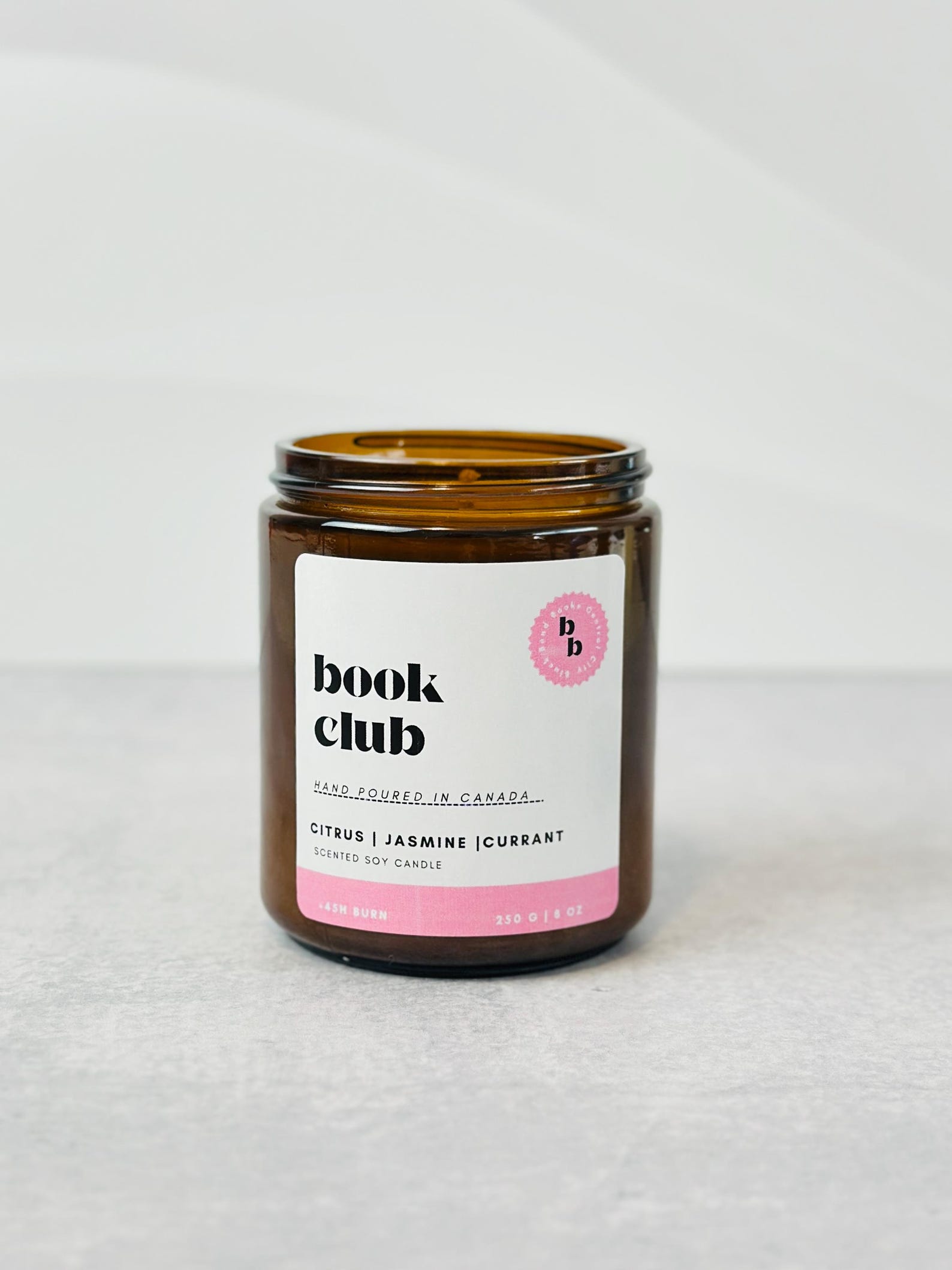 An amber candle is set against a light grey background. The label on it says "book club" in black, bold serif font and is spelt in all lower case letters. The label has pastel pink detailing.